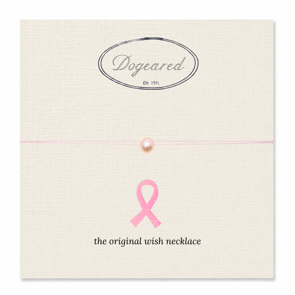 A pink wish necklace with a single pearl bead on a thin pink string, displayed on a cream-colored textured card. The card features the 'Dogeared' brand logo at the top and a pink ribbon graphic with the text 'the original wish necklace' at the bottom.