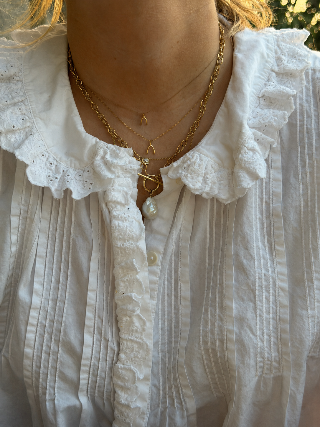 A close-up of a person wearing layered gold necklaces, including a heart-shaped pendant and a pearl drop, over a white ruffled blouse.