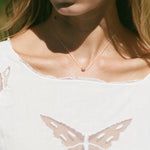 A close-up lifestyle shot of a model wearing a delicate silver chain necklace with a small, round rose gold bead pendant. The model is wearing a white top with a butterfly design on the front, set against a softly blurred green background.