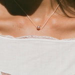 A delicate necklace with a single small pearl pendant worn by a model. The model is wearing a white lace-trimmed top with butterfly cutouts under warm, direct sunlight.