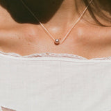 A delicate necklace with a single small pearl pendant worn by a model. The model is wearing a white lace-trimmed top with butterfly cutouts under warm, direct sunlight.