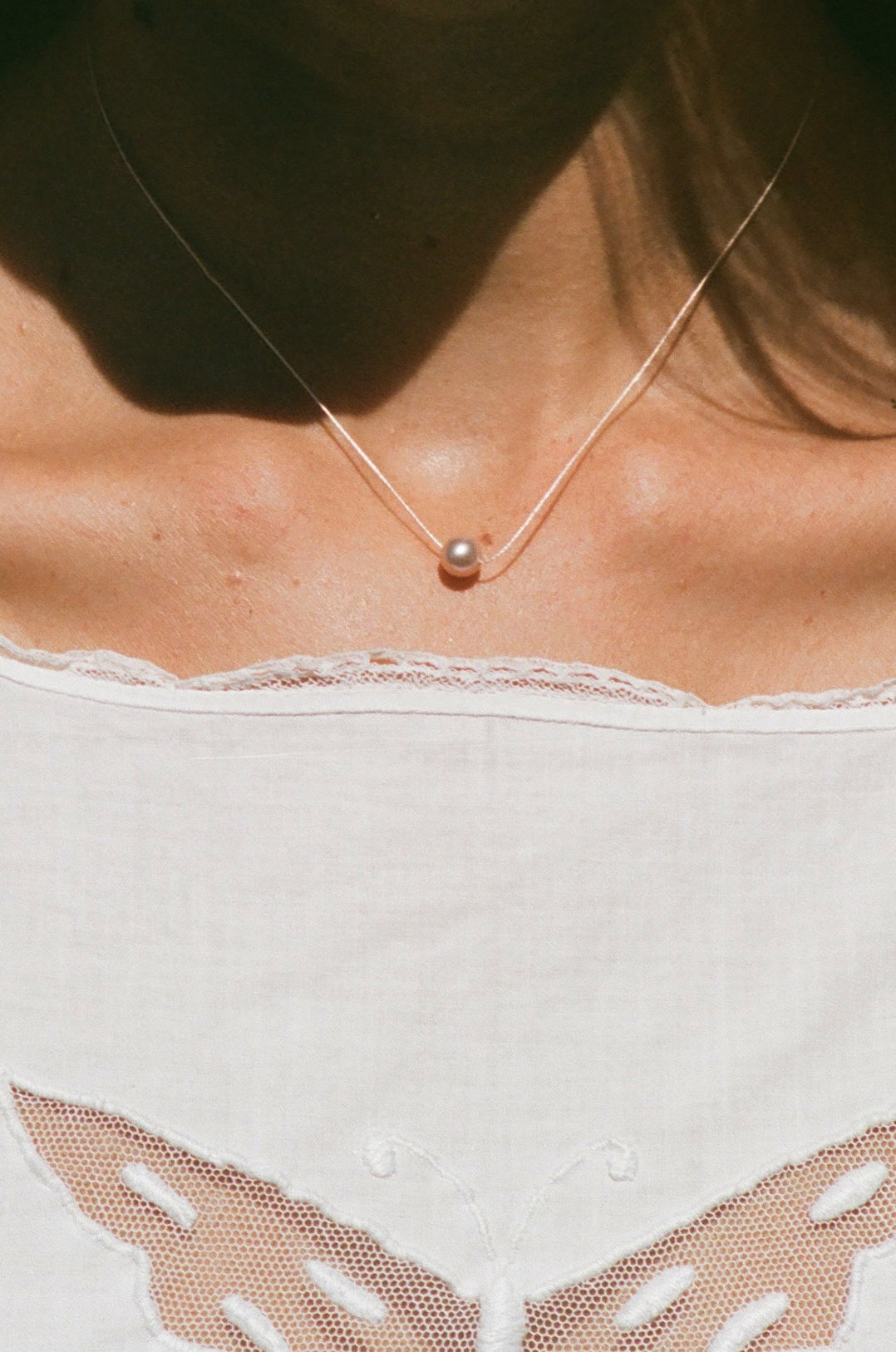 A delicate necklace with a single small pearl pendant worn by a model. The model is wearing a white lace-trimmed top with butterfly cutouts under warm, direct sunlight.