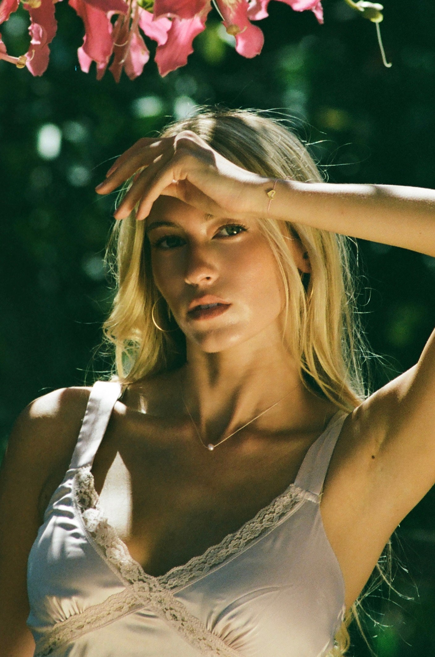 A blonde woman outdoors wearing a white lace-trim camisole top, with pink flowers hanging at the top of the frame and a blurred green natural background.
